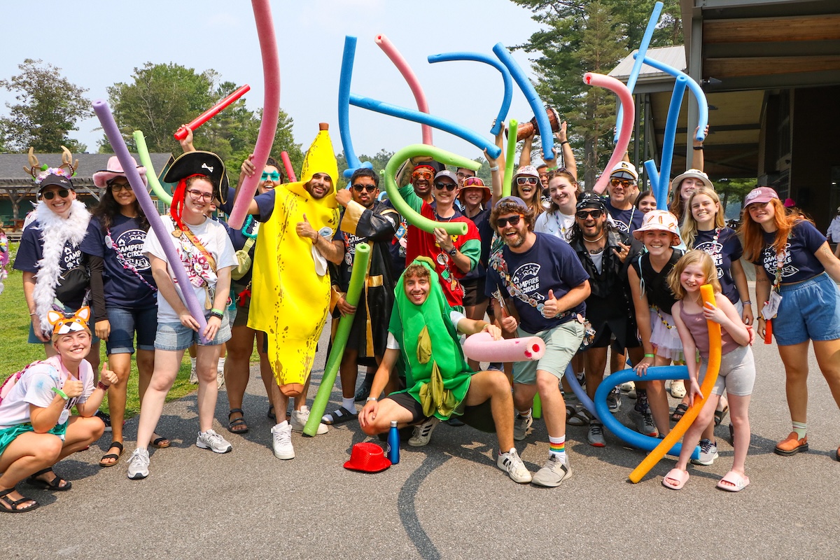 volunteers and staff in costume, waving pool noodles on a sunny day at camp